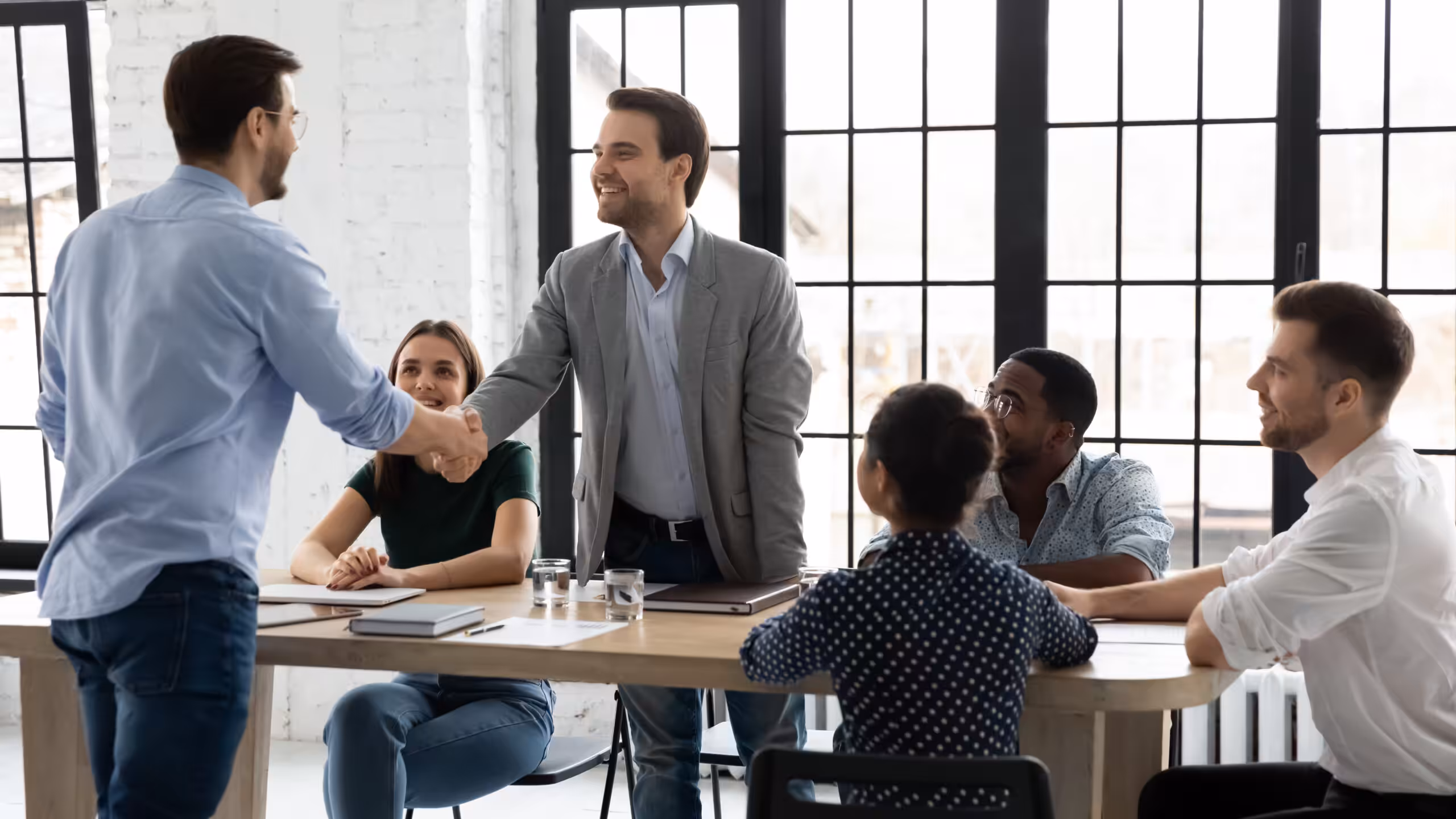 Five young professionals at a maple table with two men standing and shaking hands